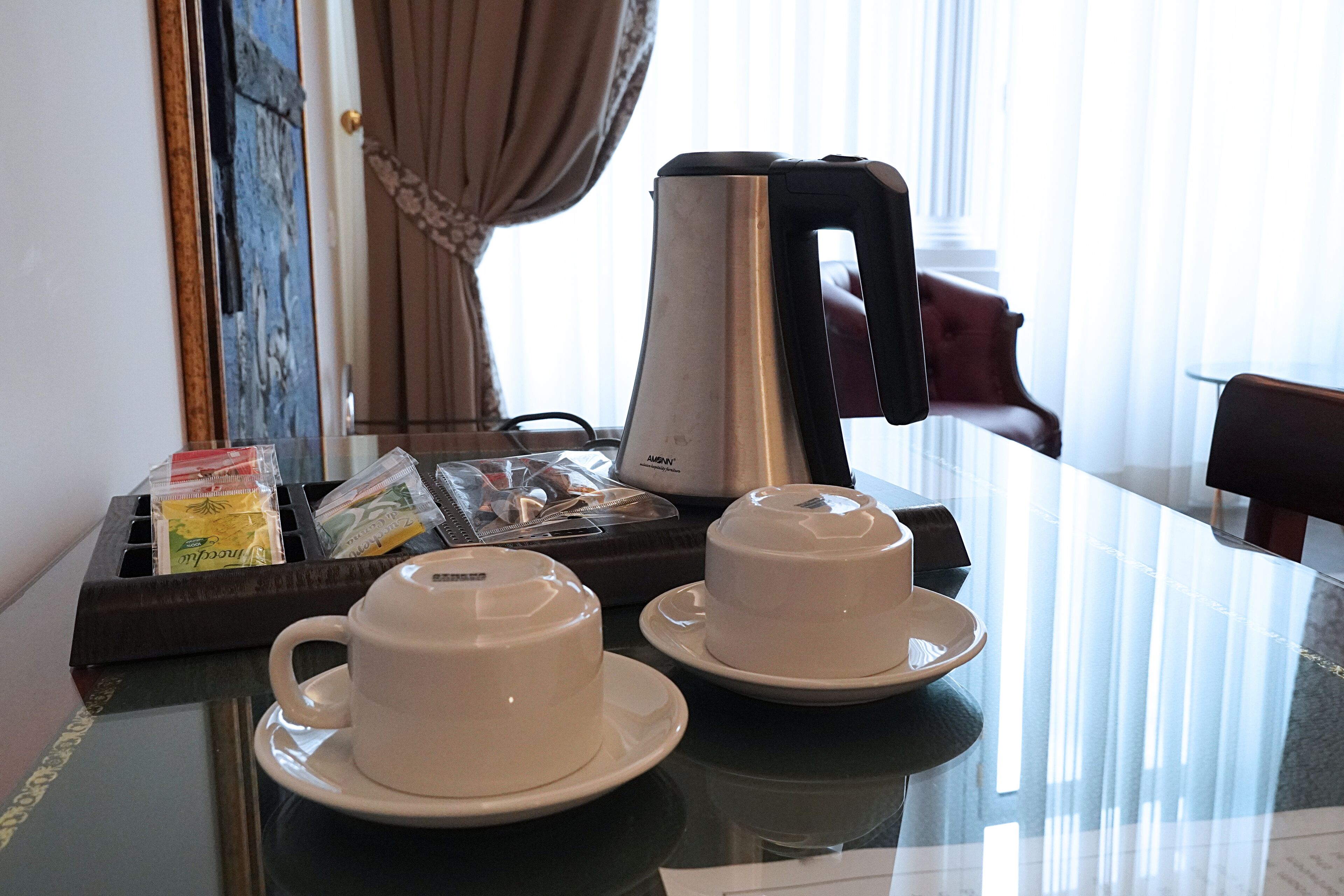A hotel room with a dining table set with two white cups and saucers, a kettle, and a tray with packets of tea and snacks.