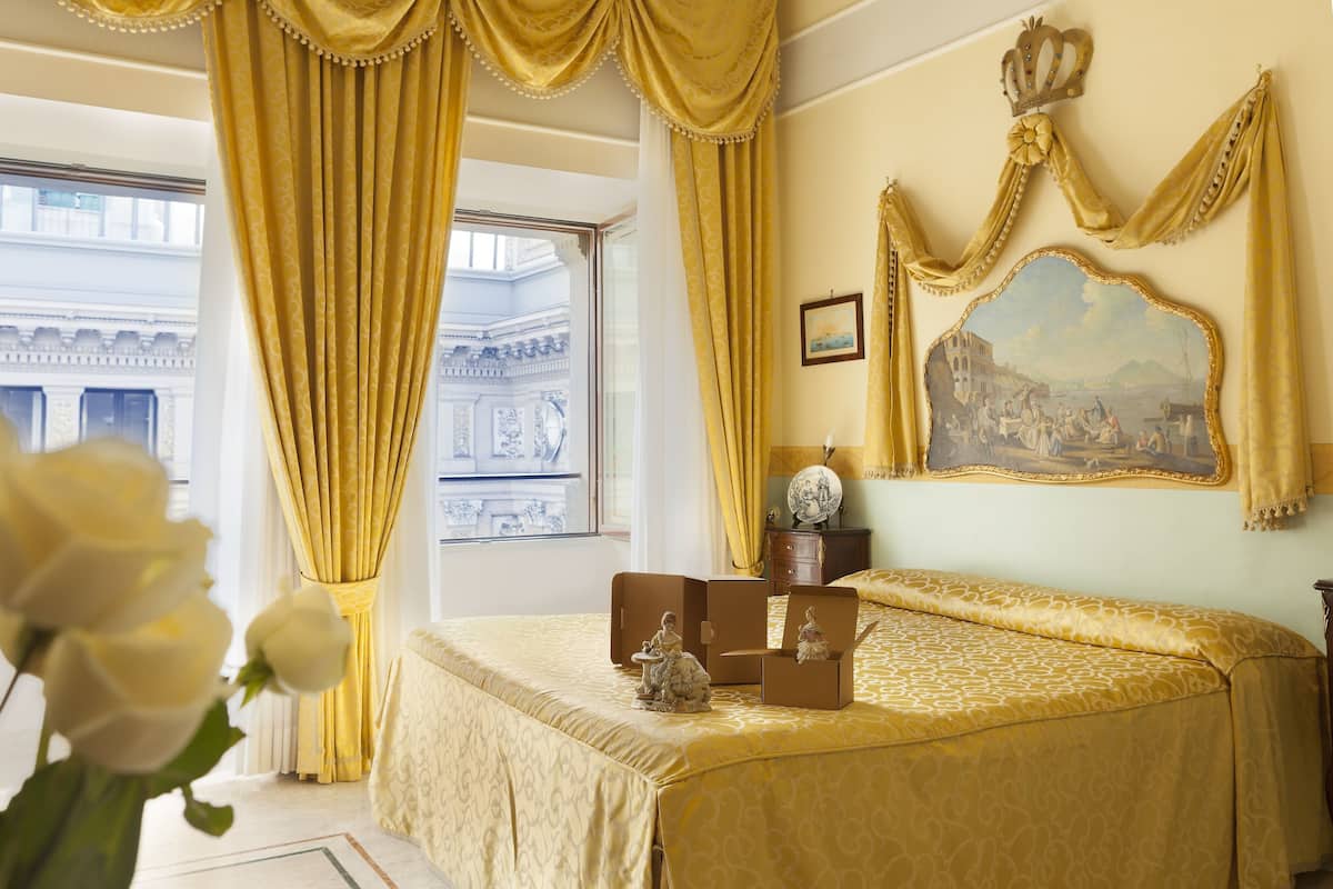 A room with a bed, a large window with curtains, a painting, and a vase with flowers.