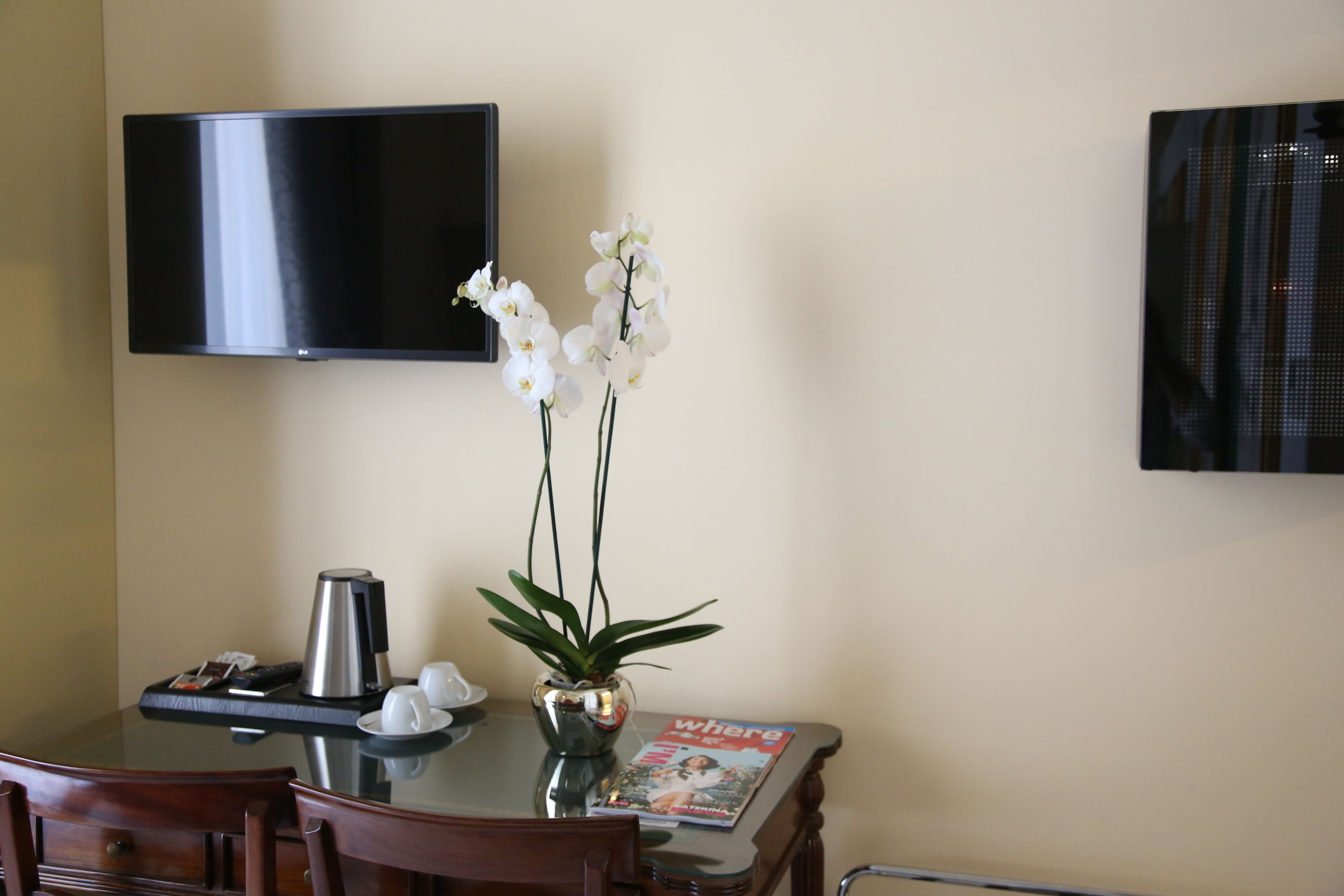 A hotel room with a dining table, chairs, a coffee maker, a vase with white orchids, a magazine, and a flat-screen TV mounted on the wall.