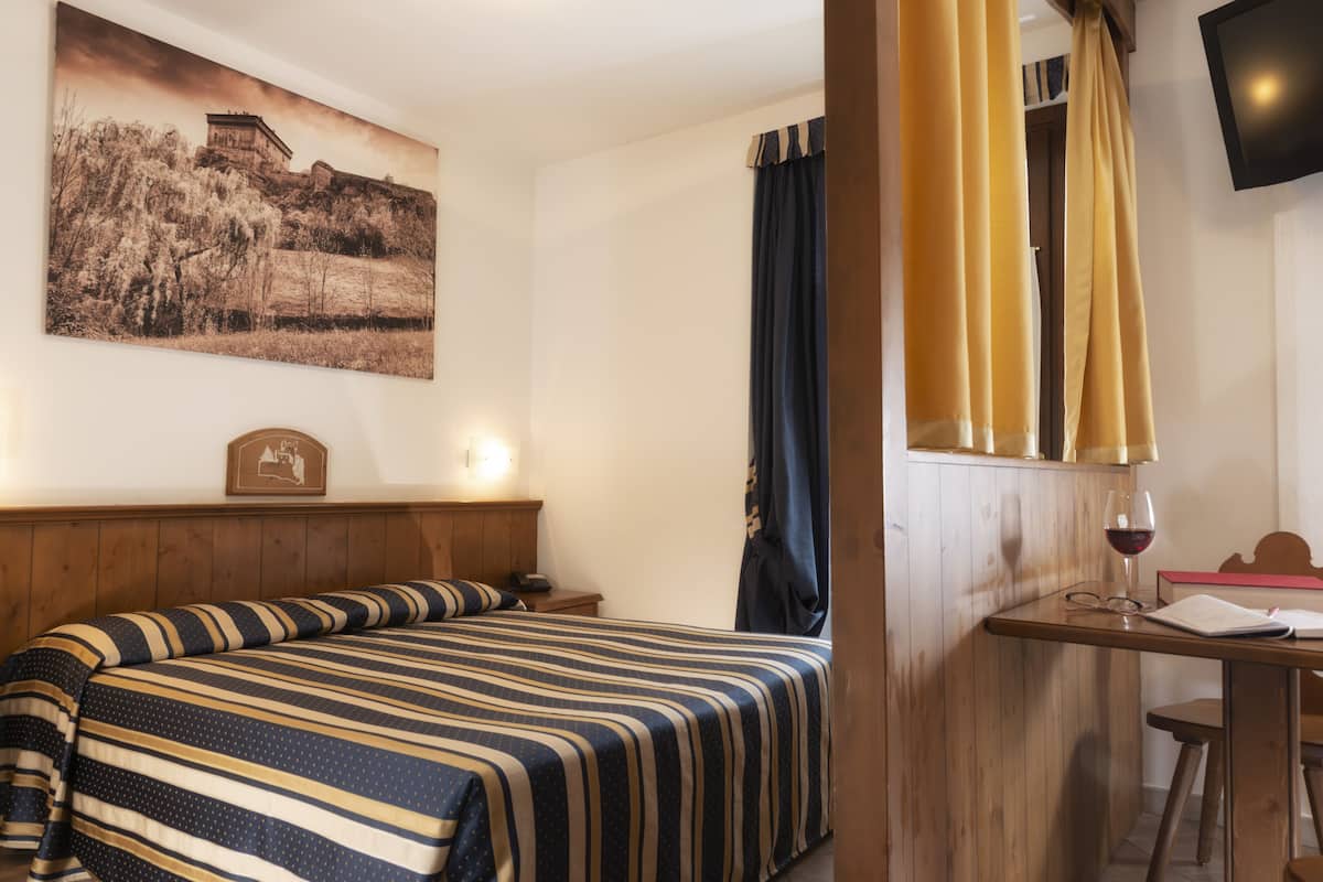 A hotel room with a bed, a wooden nightstand, a table with a book and a glass of wine, a painting on the wall, and curtains.