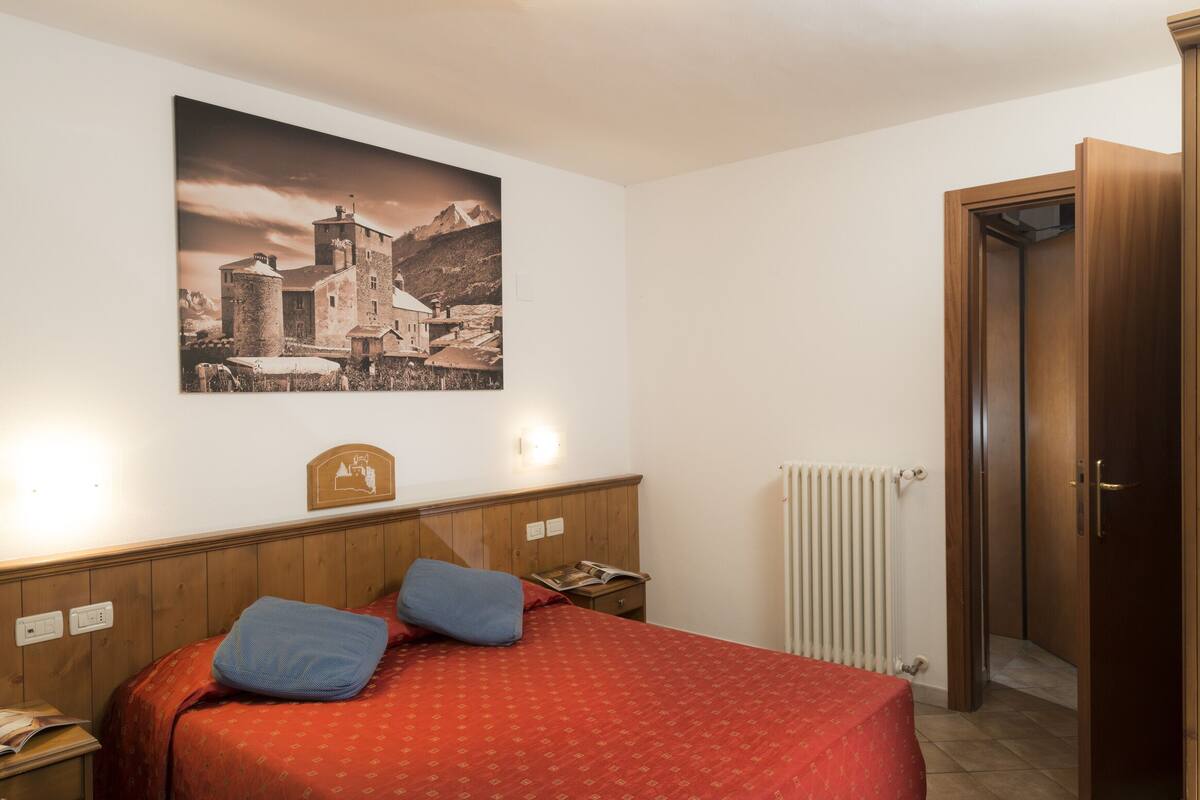 A bedroom with a wooden headboard, a bed with red bedding, a nightstand with a lamp, a framed picture on the wall, and a wooden wardrobe.