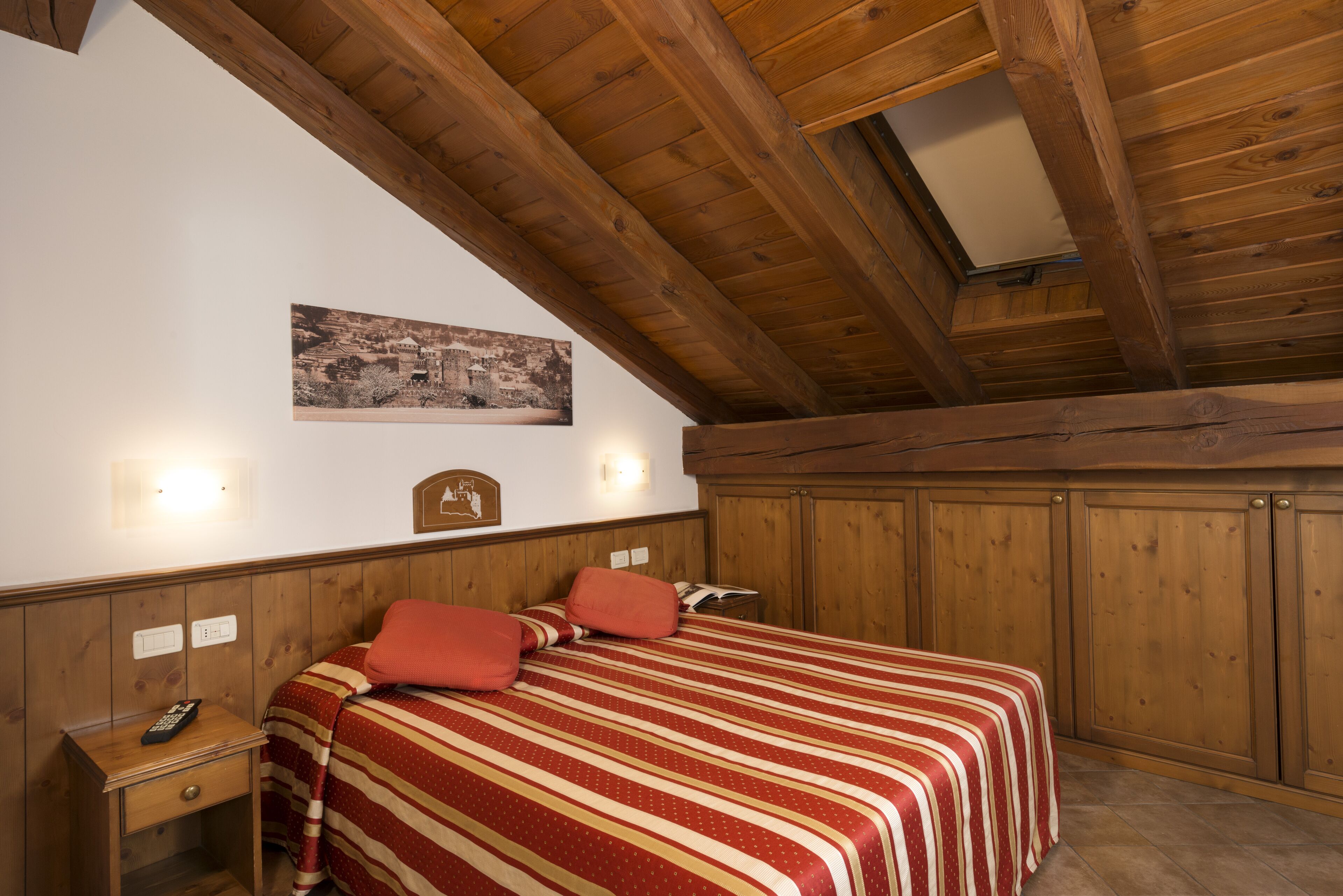 A bedroom with a wooden ceiling, a bed with red and white striped bedding, a nightstand with a remote control, and a wall-mounted light fixture.