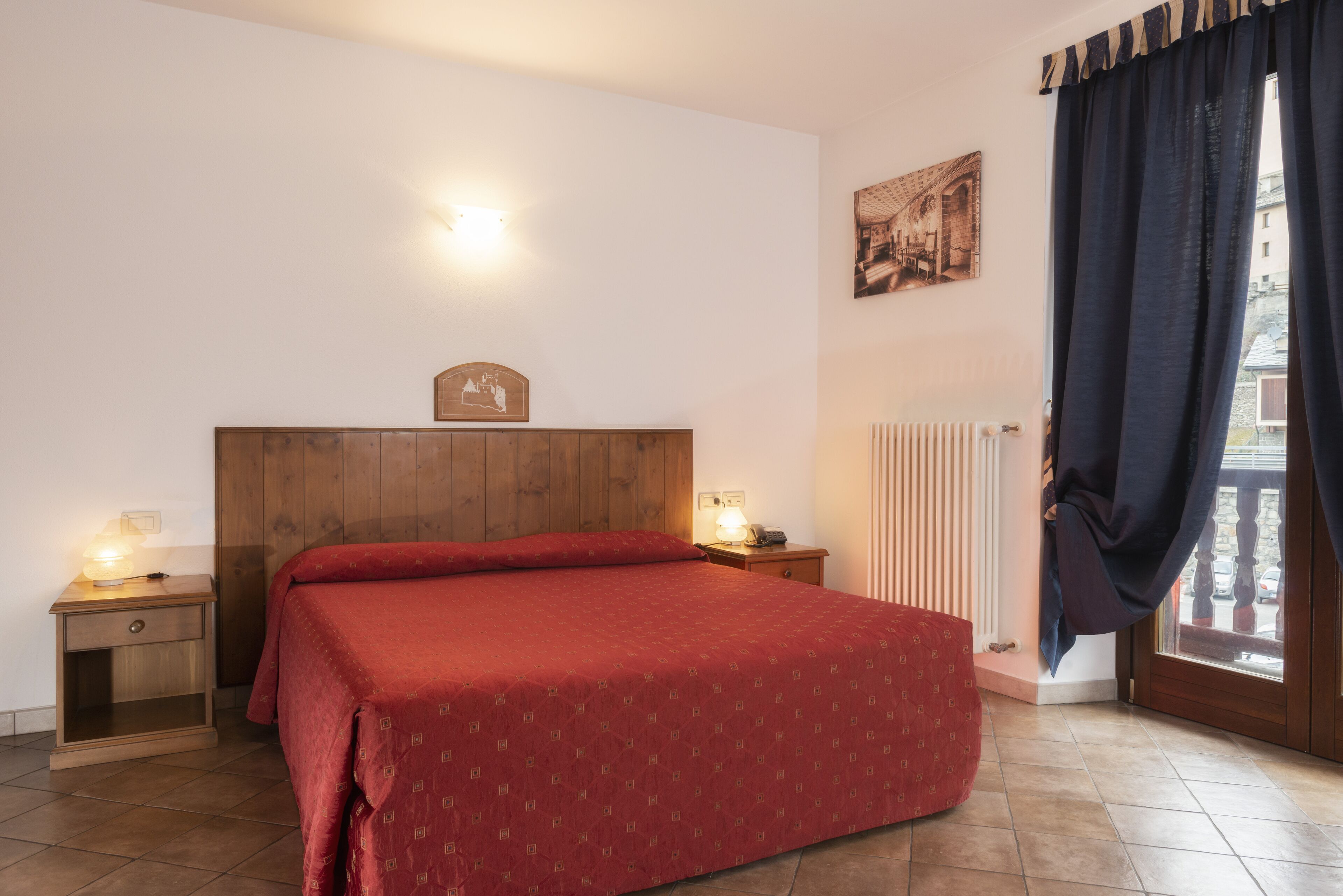 Ein Schlafzimmer mit einem hölzernen Kopfende, einer roten Bettdecke, zwei Nachttischen mit Lampen, einem Heizkörper und einem Fenster mit Vorhängen.