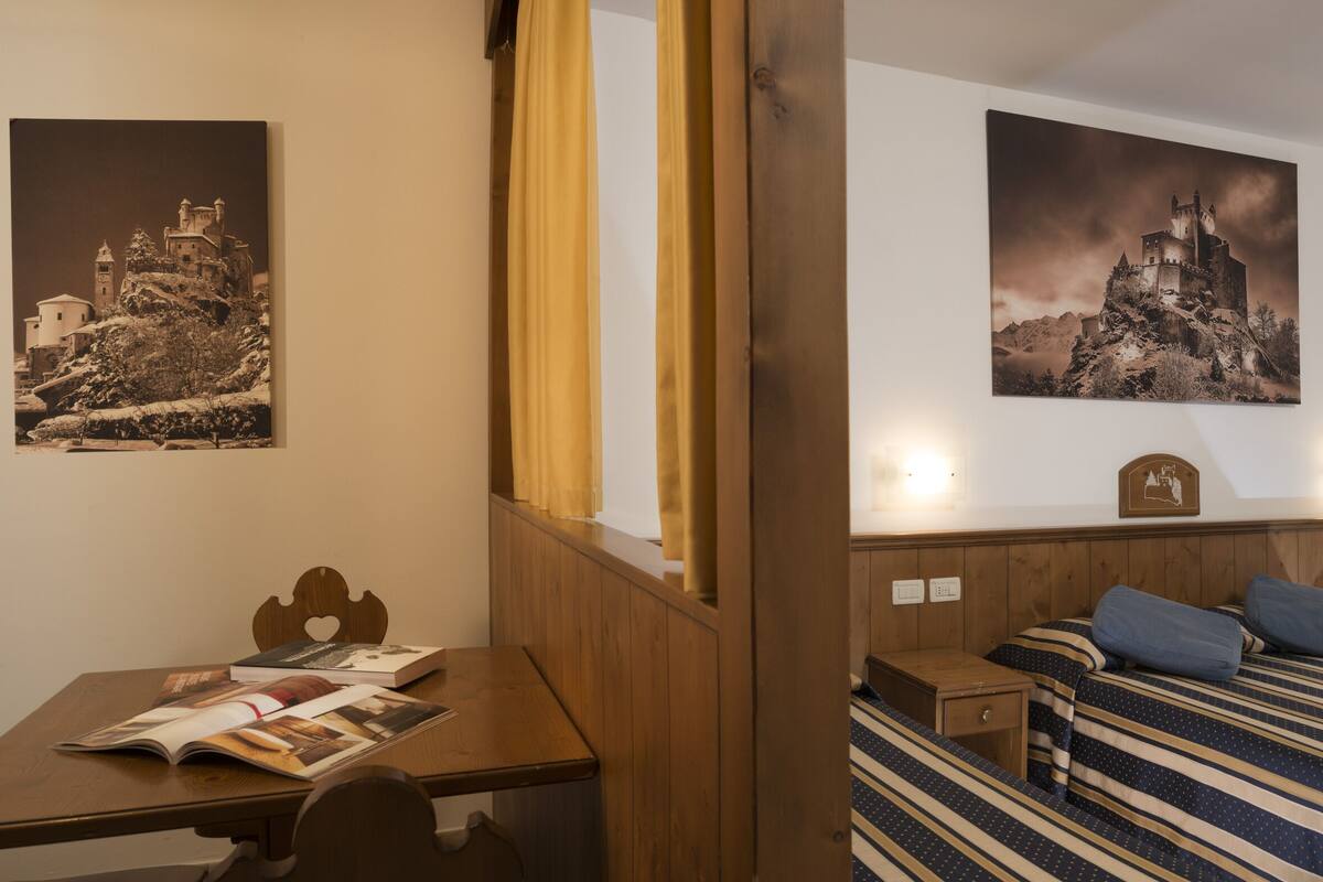 A hotel room with a wooden bed, a small bedside table, a wooden nightstand, a wooden chair, a wooden table, and two wall-mounted light fixtures.