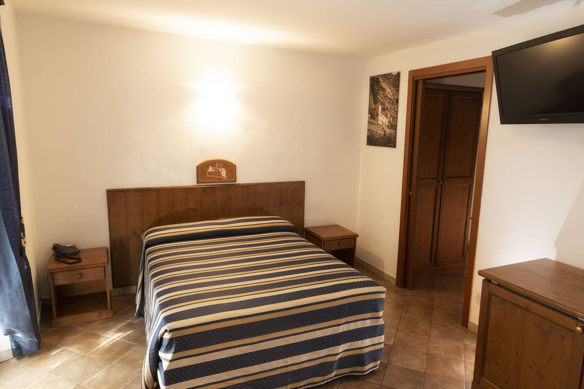 A bedroom with a bed, wooden headboard, bedside tables, a nightstand, a television mounted on the wall, and a door leading to another room.
