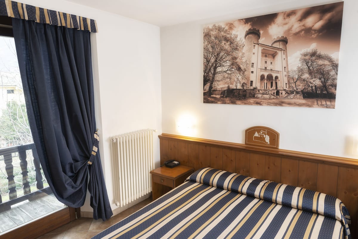 A bedroom with a bed, a nightstand, a lamp, a window with curtains, and a wall-mounted picture of a castle.