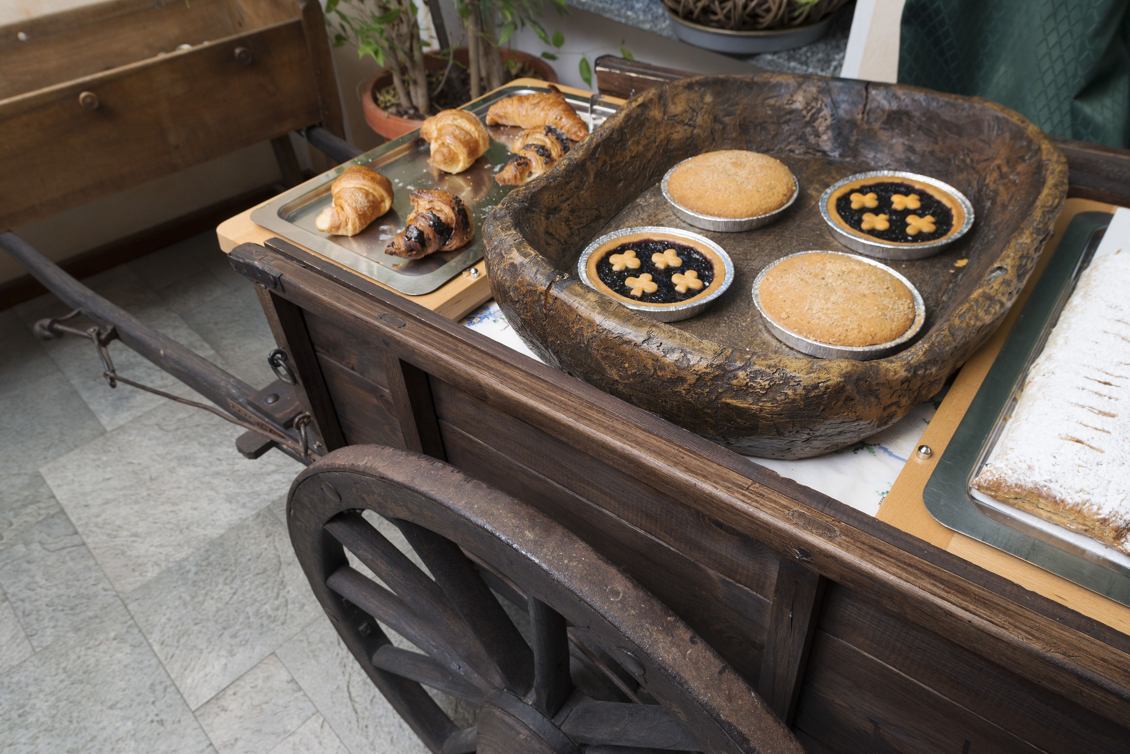 A rustic wooden cart with a tray holding croissants and three small metal pans with baked goods.