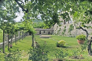 A courtyard with a stone building, a grassy lawn, and a pergola with climbing plants.