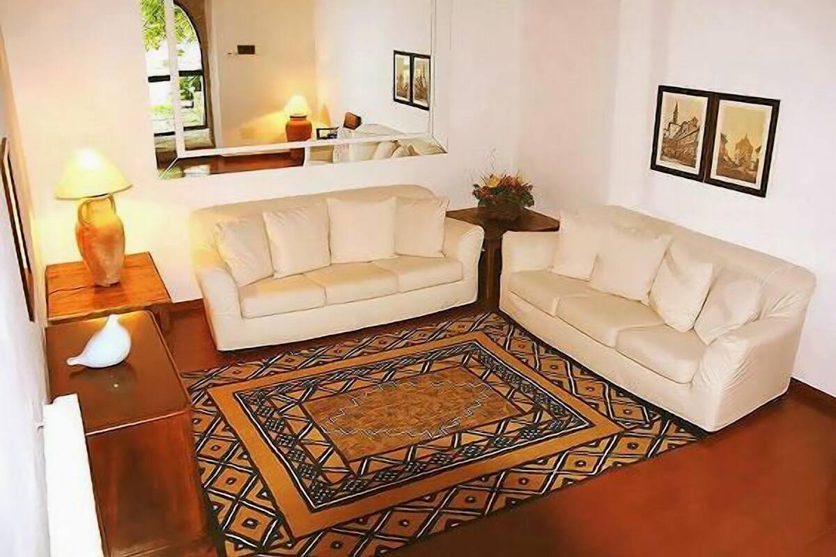 Ein Wohnzimmer mit zwei weißen Sofas, einem gemusterten Teppich, einem kleinen Tisch mit einer Lampe und gerahmten Bildern an der Wand.