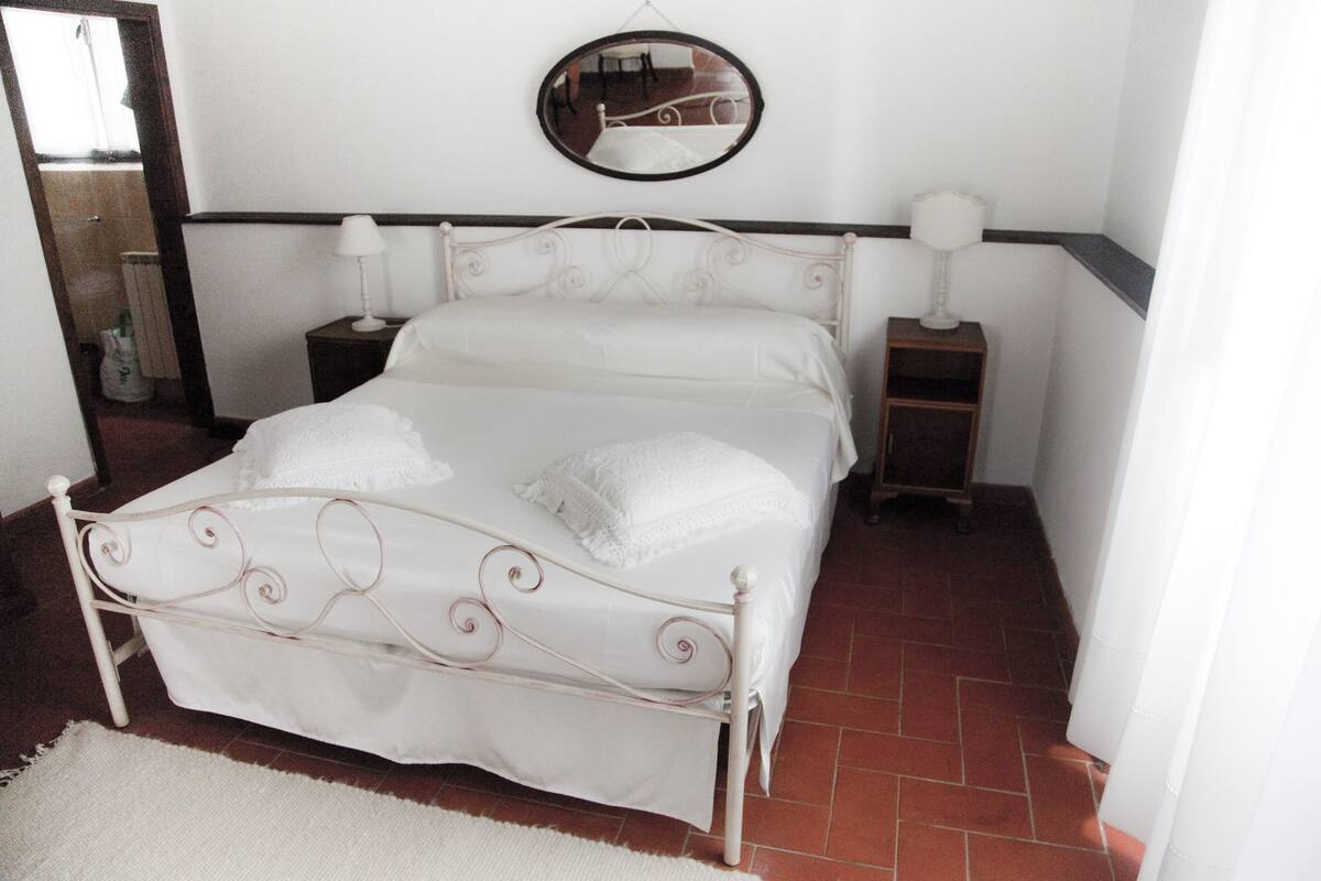 Ein weißes, metallenes Himmelbett mit einem Spiegel darüber, zwei Nachttische und ein weißer Teppich auf Fliesenboden.