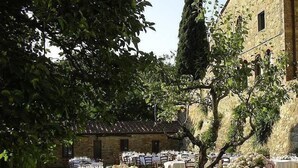An outdoor dining area with tables set for a meal, surrounded by greenery and a stone building in the background.