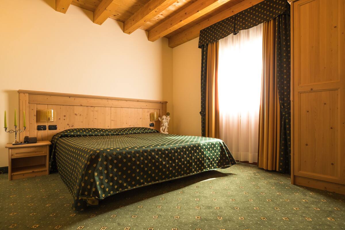 Une chambre à coucher avec une tête de lit en bois, un lit recouvert d’une couvre-lit à motifs verts, une table de chevet avec une lampe et une fenêtre avec des rideaux.