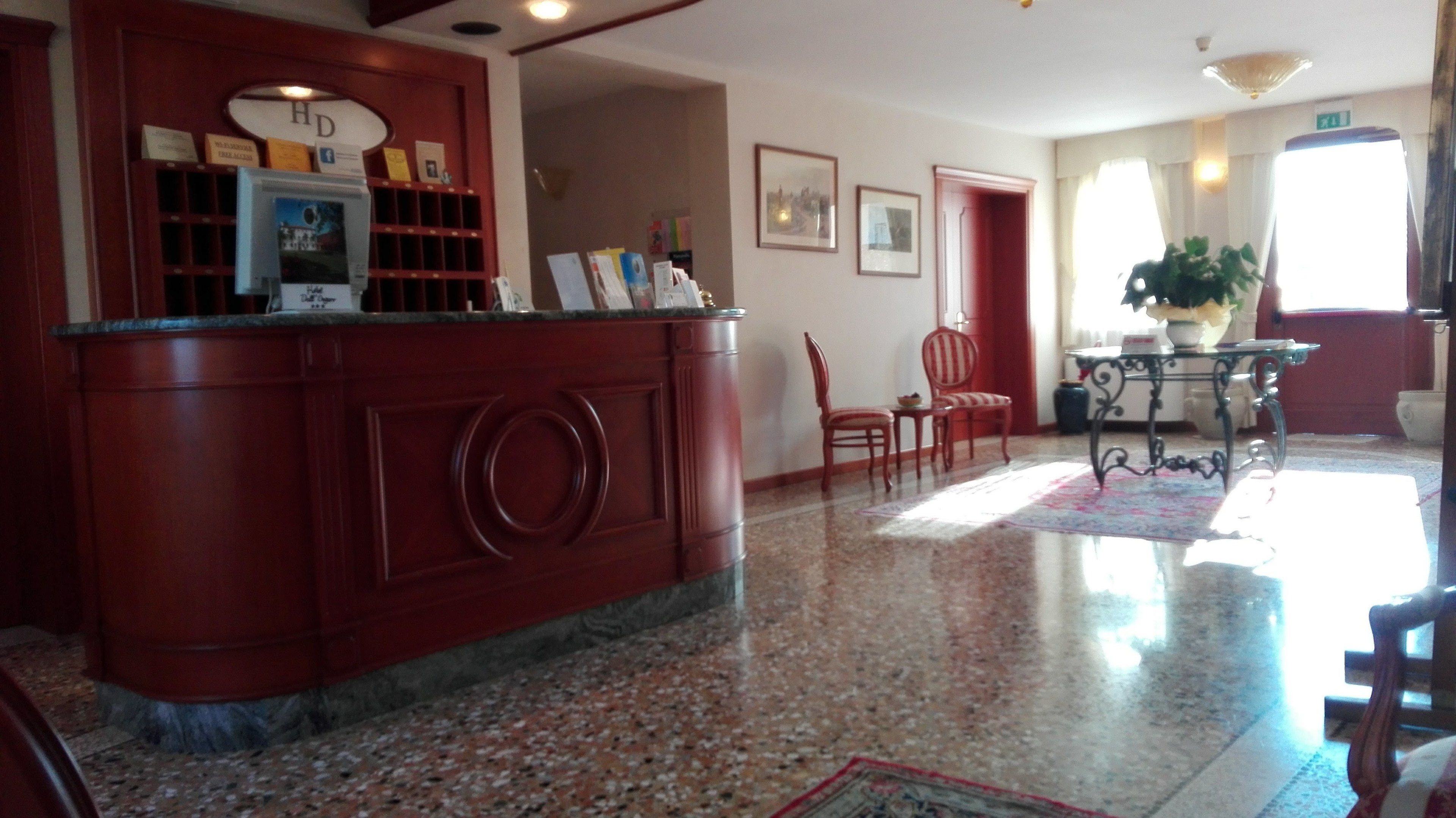 Uma área de recepção com piso de mármore, balcão de recepção, cadeiras, uma mesa com um vaso, quadros nas paredes e uma porta.