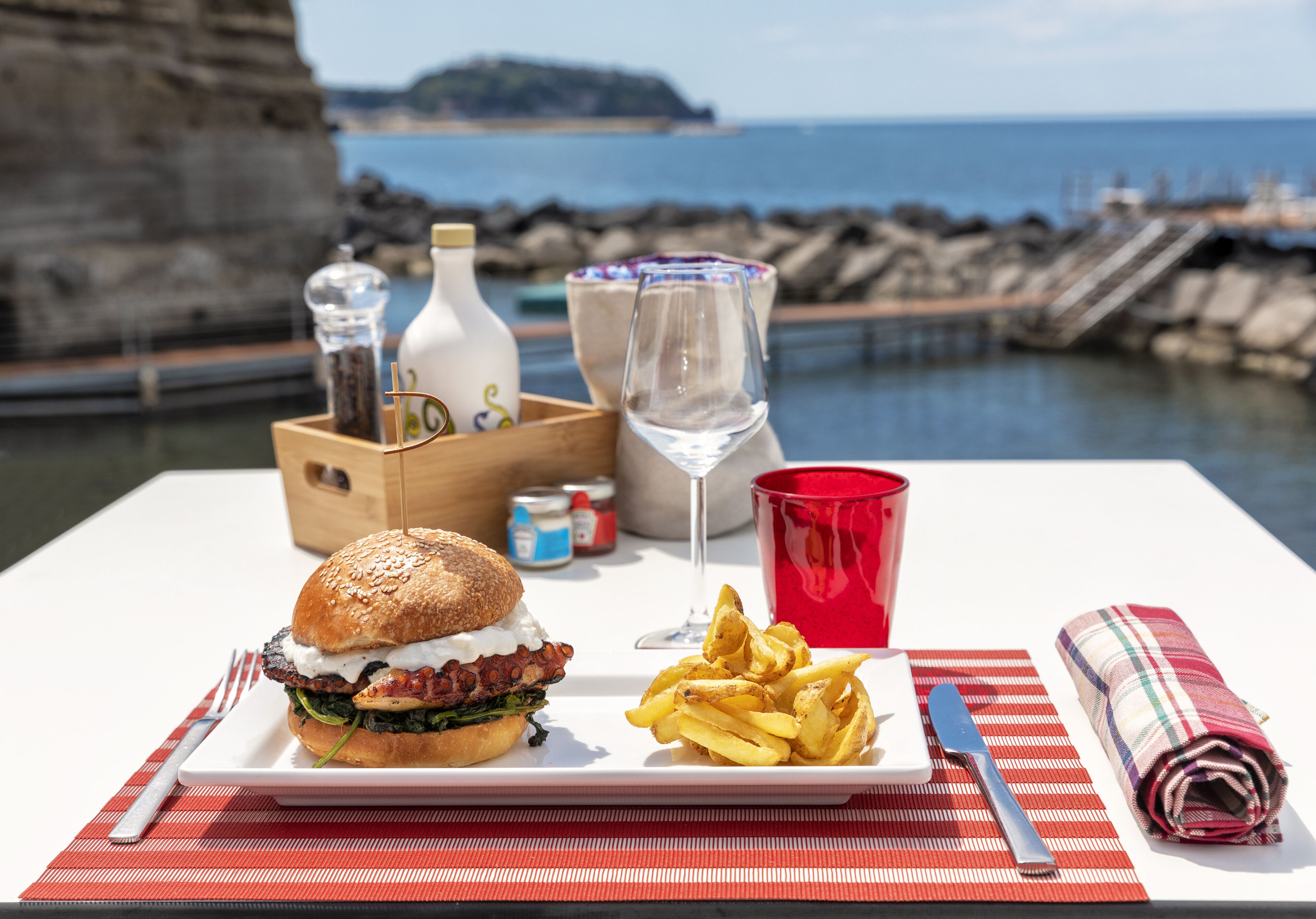 A meal with a burger, fries, and a glass of wine on a table with a view of the sea and cliffs.