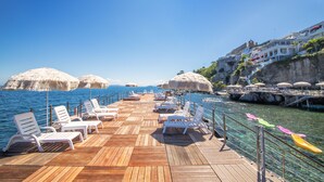Une terrasse en bois, agrémentée de chaises longues blanches et de parasols en chaume, offrant une vue imprenable sur une mer bleue et une côte rocheuse.