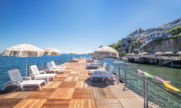 Una terraza de madera con sillones de descanso blancos y sombrillas de paja, con vista a un mar azul y una costa rocosa.