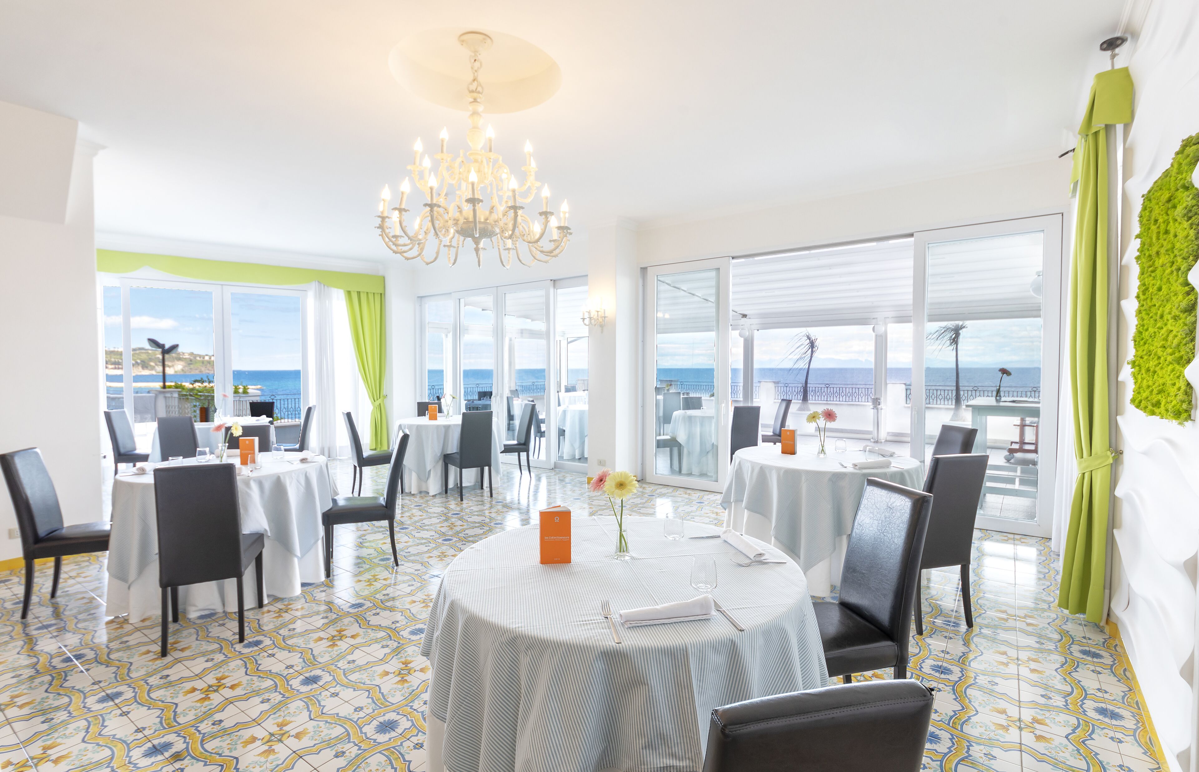 Un espace repas avec des tables rondes, des chaises, un lustre et une vue sur la mer.
