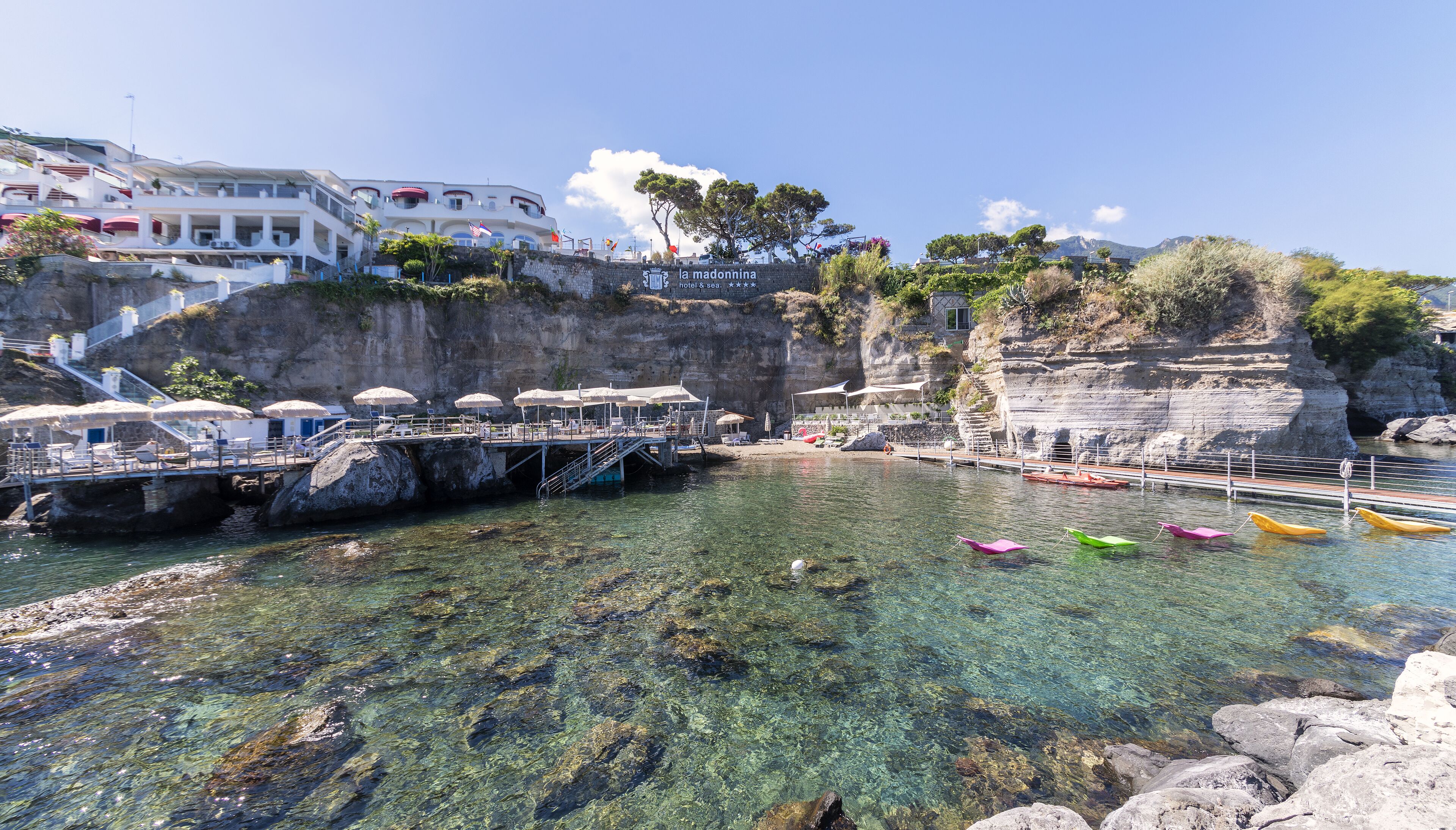 A coastal hotel with a clear view of the sea, a rocky shoreline, and a pier with umbrellas.