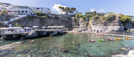 A coastal hotel with a clear view of the sea, a rocky shoreline, and a pier with umbrellas.