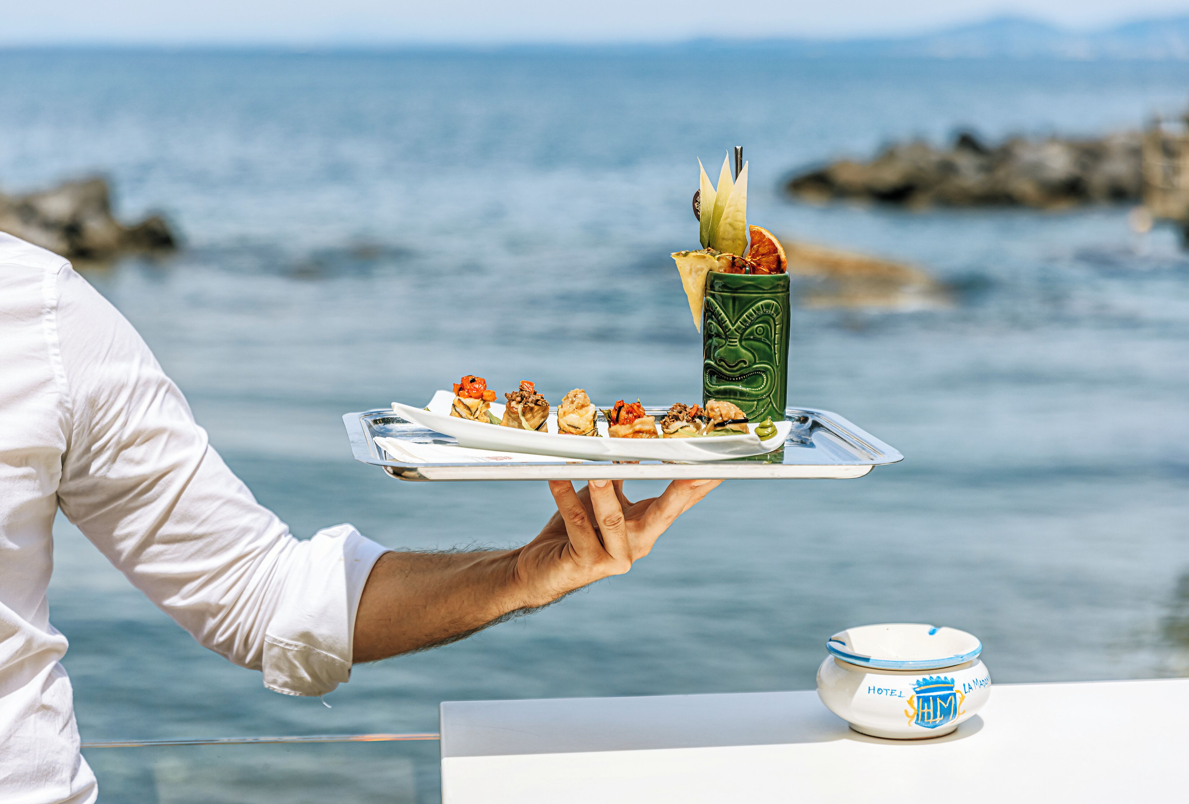 Una persona sosté una safata amb un plat i una beguda, amb un cos d'aigua i roques al fons.
