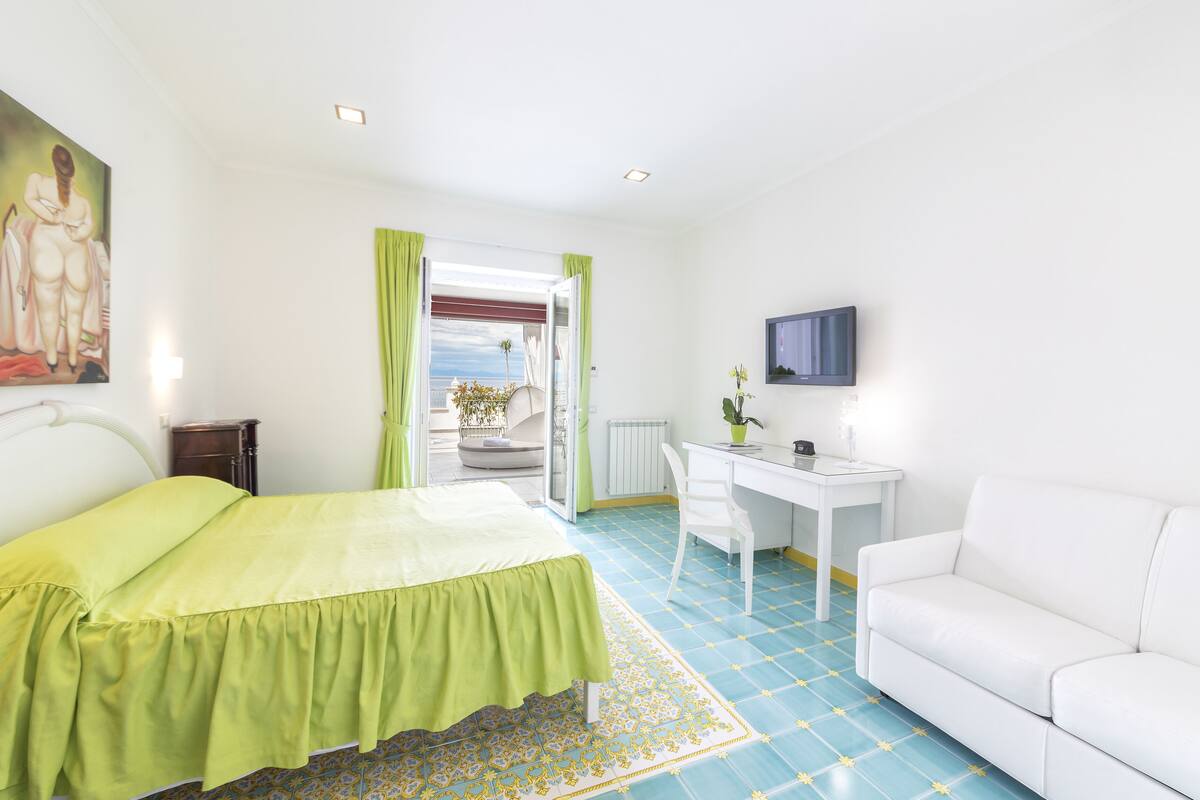 Ein Schlafzimmer mit einem Bett, einem Schreibtisch, einem Stuhl, einem weißen Sofa, einem Gemälde und Blick durch eine offene Tür.