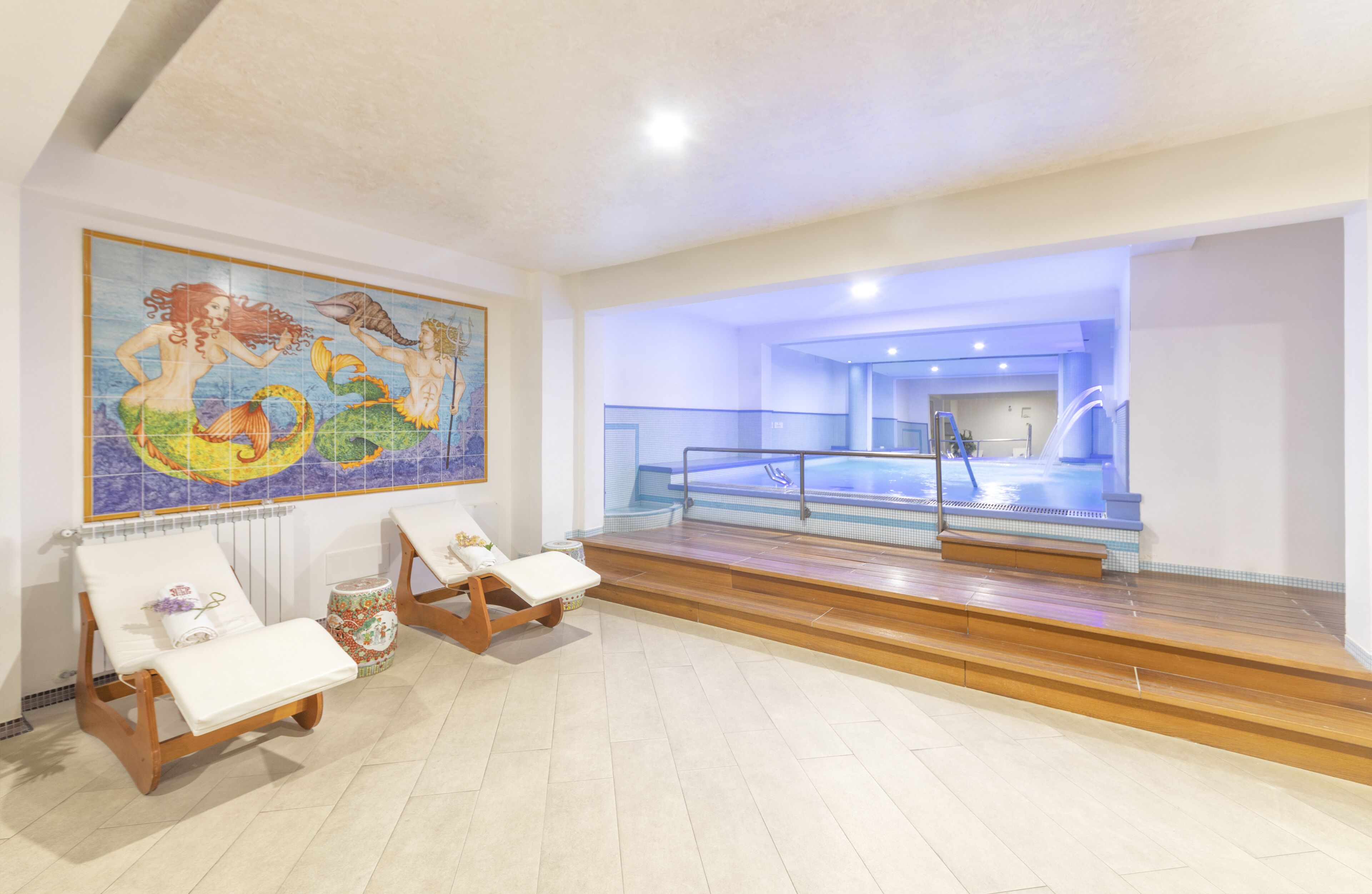 A spa area with two sun loungers, a mosaic wall, and a pool with a glass barrier.