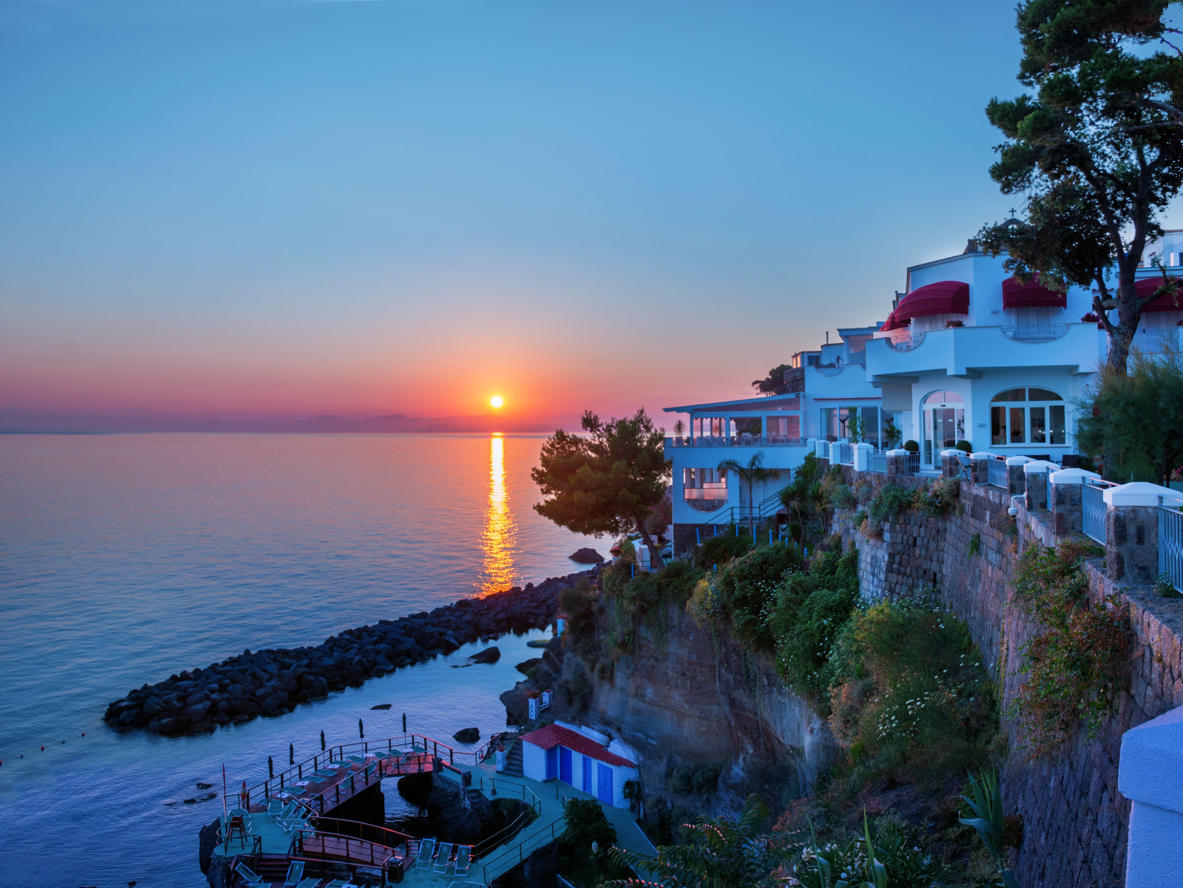 A coastal hotel with a view of the sunset over the sea, a pool area, and a cliffside location.
