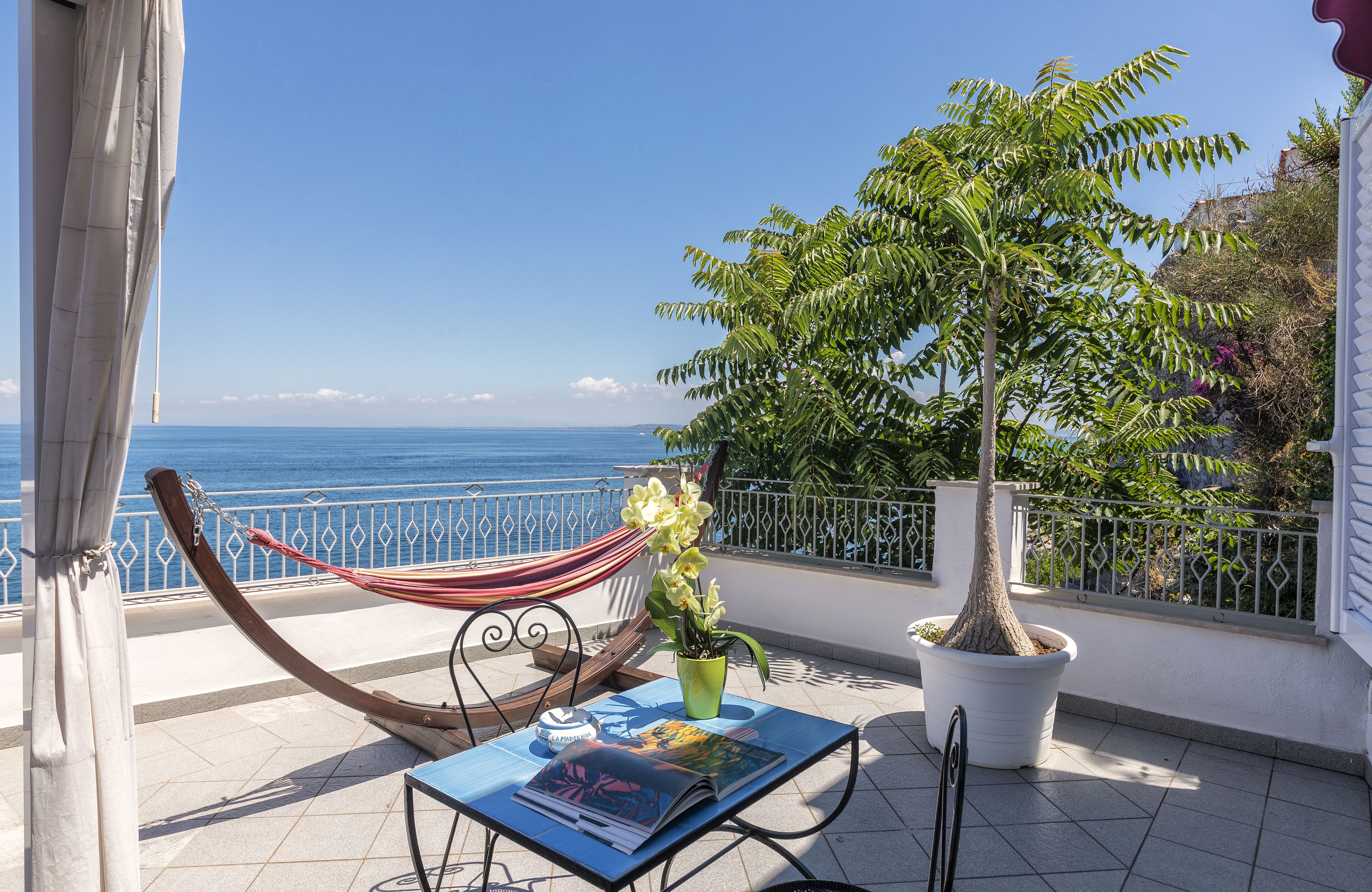A balcony with a hammock, a table with magazines, a potted plant, and a view of the sea.