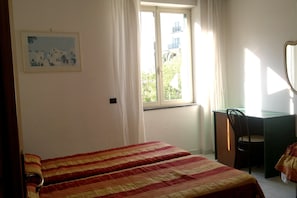 Una habitación con cama, escritorio y silla. Tiene una ventana con cortinas, un cuadro en la pared y una puerta.