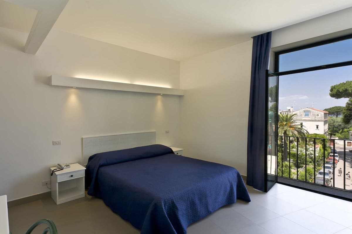 A modern bedroom with a large bed, bedside table, and a view of the street through a balcony.