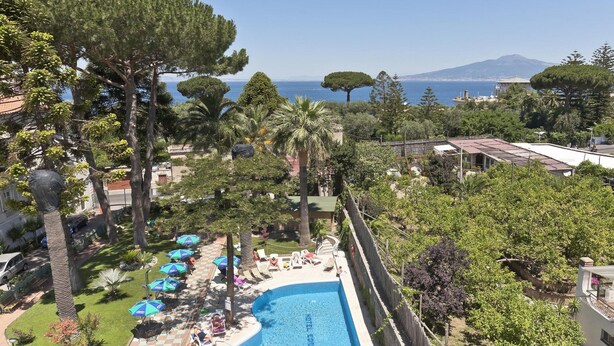 A pool area with lounge chairs and umbrellas, surrounded by trees and a view of the sea and a mountain. - Hotel Eden (Sorrento)