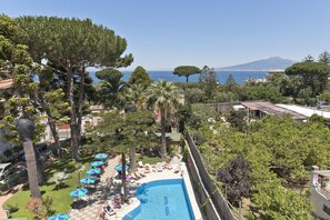 A pool area with lounge chairs and umbrellas, surrounded by trees and a view of the sea and a mountain. - Hotel Eden (Sorrento)
