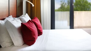 Una cama bien hecha con almohadas blancas y rojas, un cabecero de madera y vistas al exterior con vegetación.