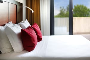 Una cama bien hecha con almohadas blancas y rojas, un cabecero de madera y vistas al exterior con vegetación.