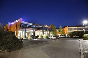 Entrada de hotel con diseño moderno, zona de estacionamiento techada y varios coches aparcados.