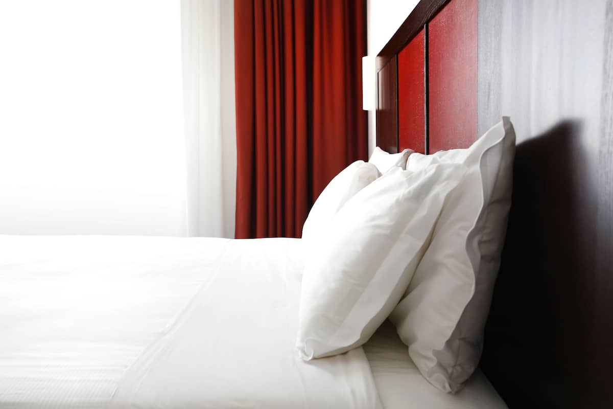 A neatly made bed with white linens and pillows, a wooden headboard, and red curtains.
