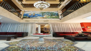 A spacious lobby with a large fountain, red sofas, a colorful patterned rug, and a mural on the wall. - Hotel Le Caspien (Marrakech)