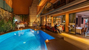 An indoor swimming pool with tables and chairs around it, and a restaurant area with patrons dining. - Hotel Le Caspien (Marrakech)