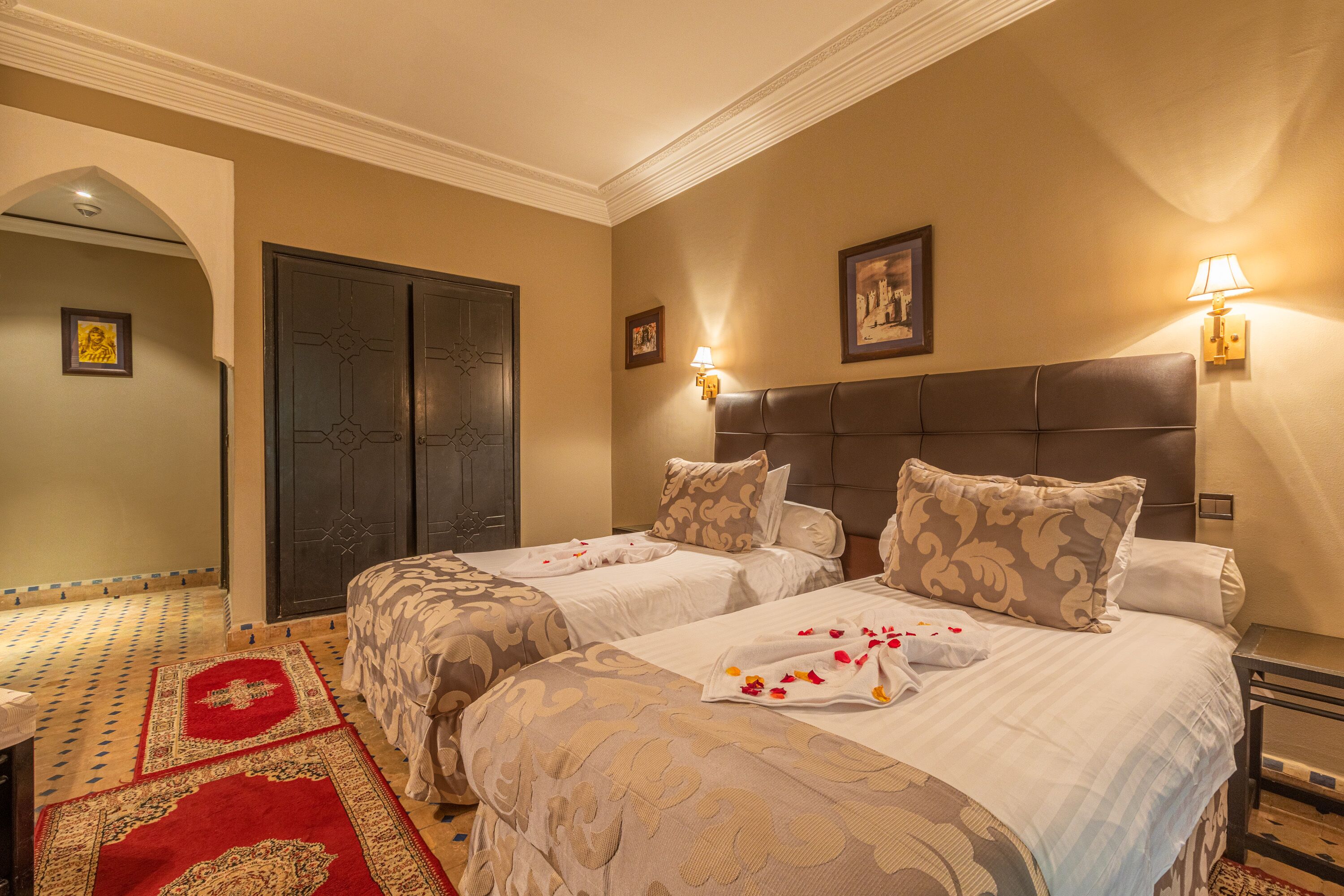 A hotel room with two beds, a large dark wooden door, a red and beige patterned rug, and wall-mounted lamps.