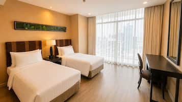 Habitación de hotel con dos camas, un escritorio, una silla, un ventanal con cortinas y vista a la ciudad.