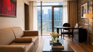 Ein modernes Hotelzimmer mit Sofa, Schreibtisch, Couchtisch mit Büchern, einer Vase mit Blumen und einem großen Fenster mit Blick auf die Stadt.