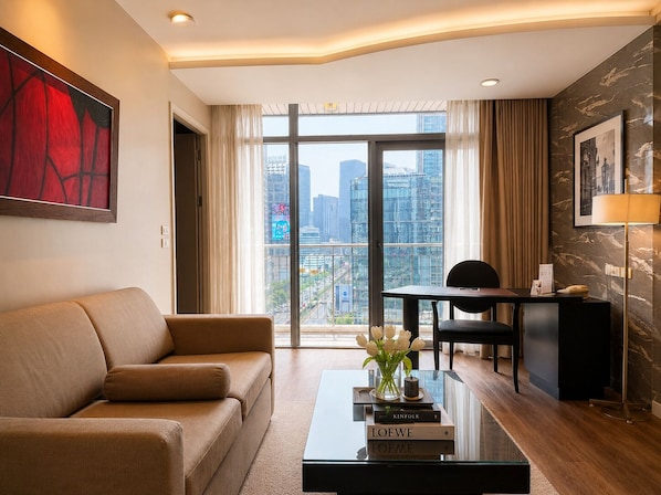 A modern hotel room with a sofa, a desk, a coffee table with books, a vase with flowers, and a large window with a city view.
