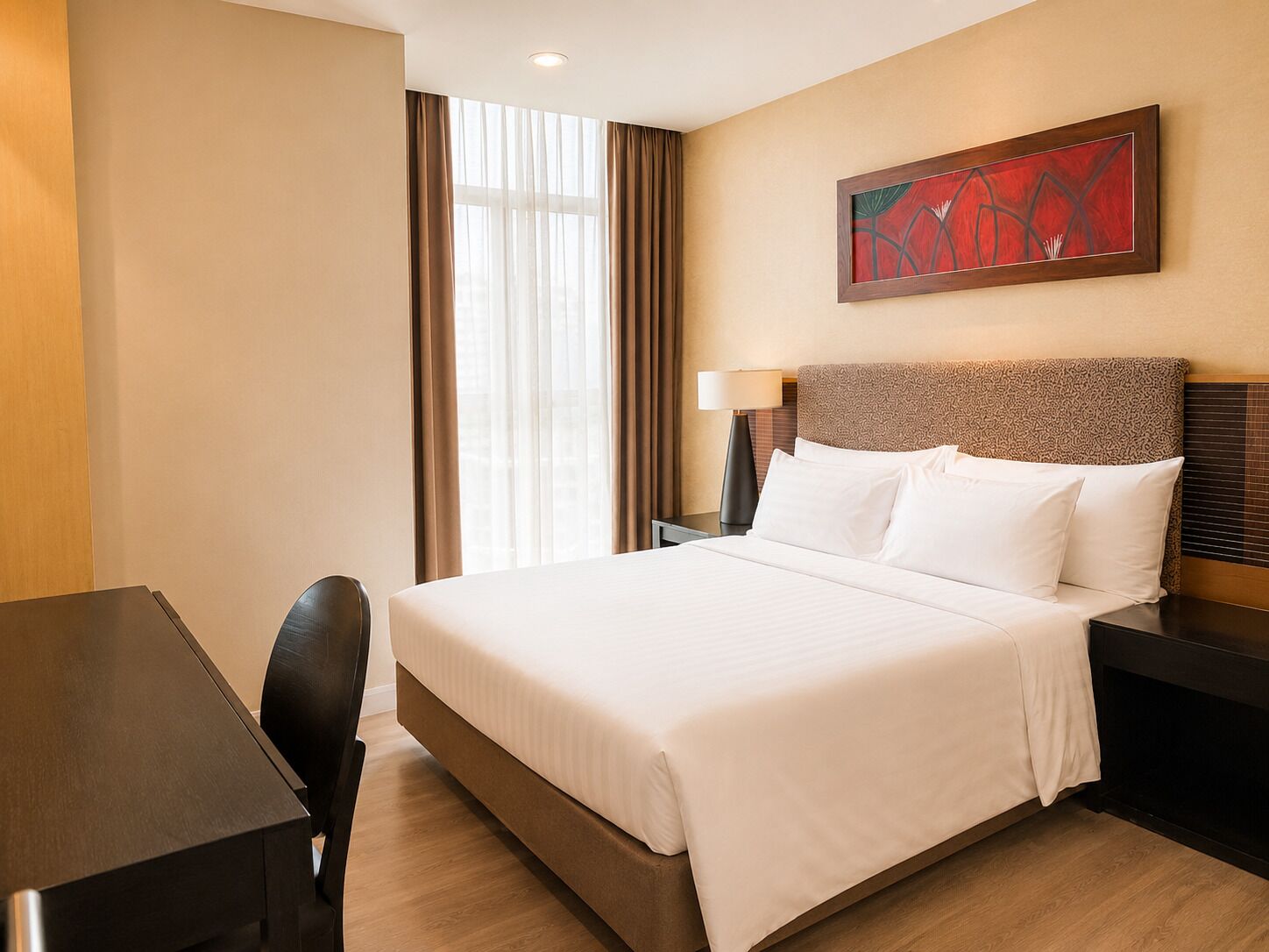 A hotel room with a bed, a desk, a chair, a lamp, and a painting on the wall.