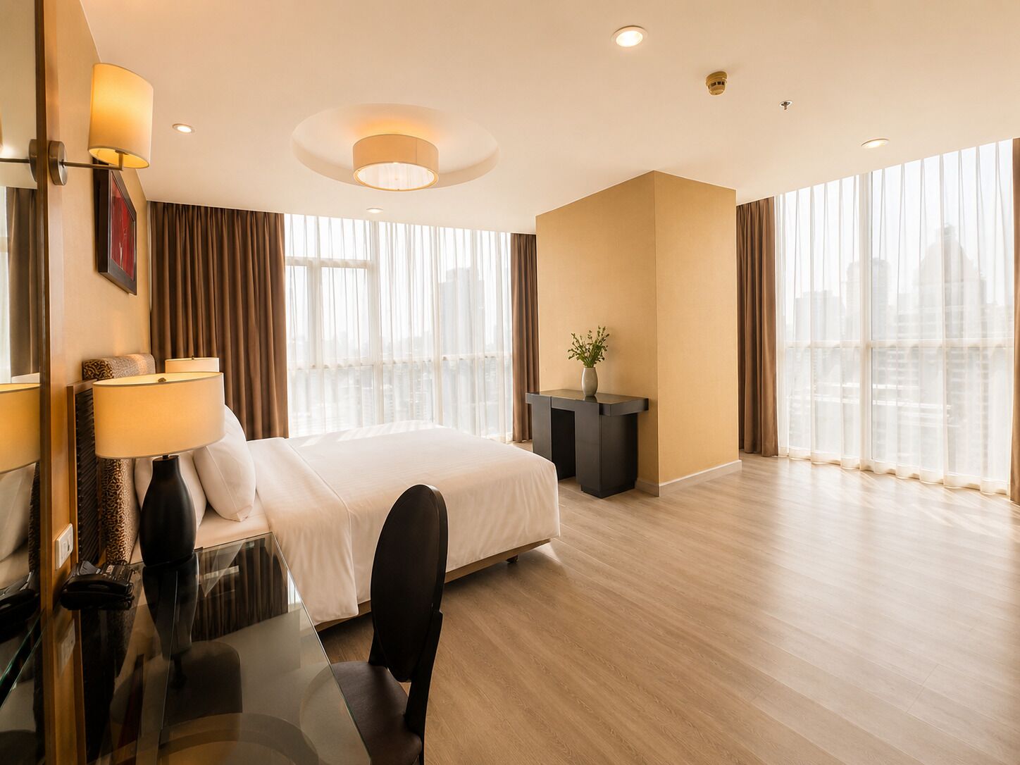 Ein Hotelzimmer mit einem Bett, einem Schreibtisch mit Lampe, einem Sessel, einem kleinen Tisch mit Pflanze und großen Fenstern mit Vorhängen.