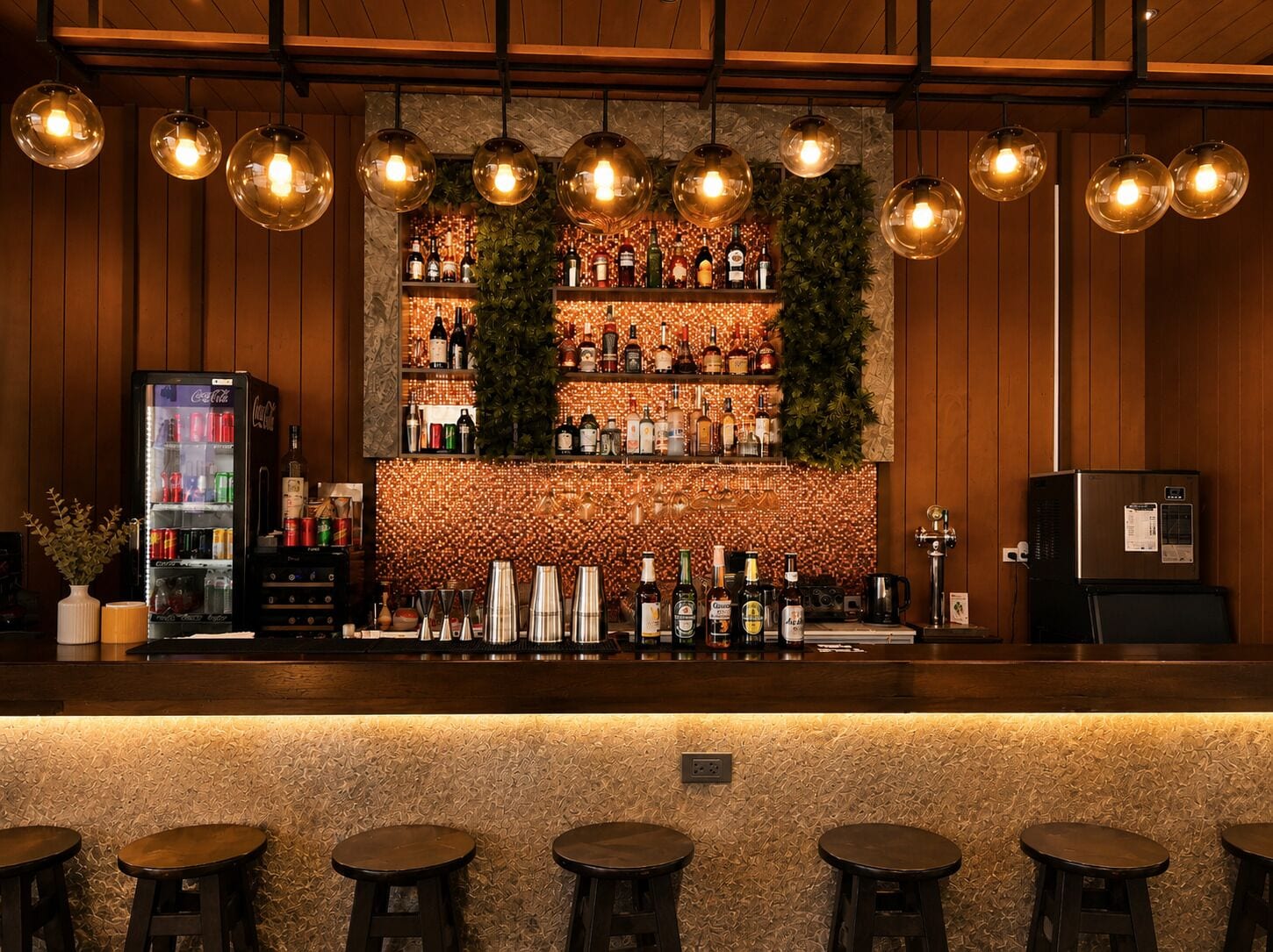 A bar area with a wooden counter, shelves of bottles, and hanging light fixtures.