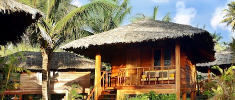 A wooden bungalow with a thatched roof, surrounded by lush greenery and palm trees.