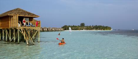 A wooden overwater bungalow with a staircase leading to a deck, clear turquoise water, and a distant island with vegetation.