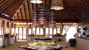A buffet setup with multiple serving stations, a high ceiling with wooden beams, and large windows allowing natural light.
