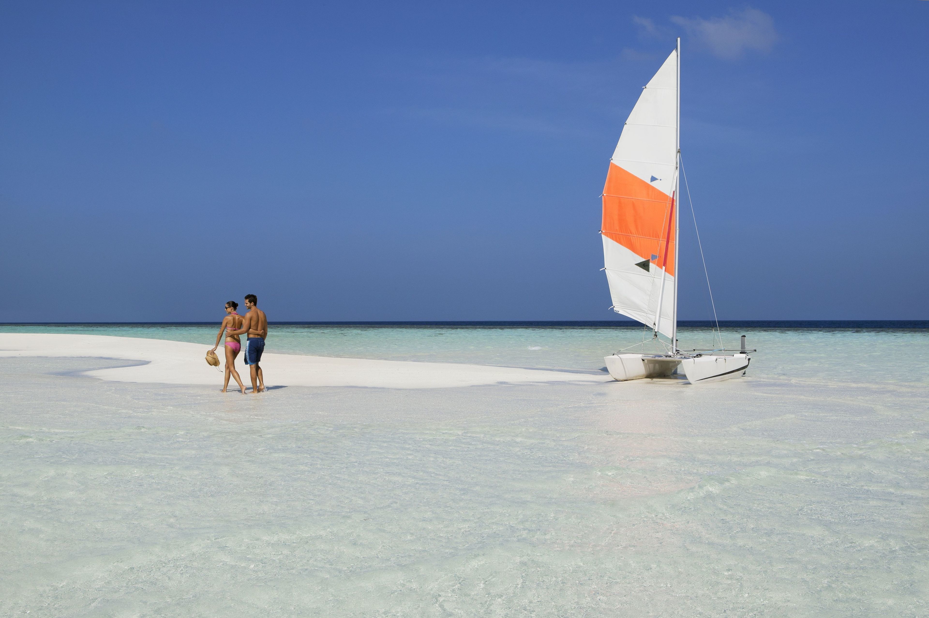 A couple walking on a sandy beach with a sailboat in the water.