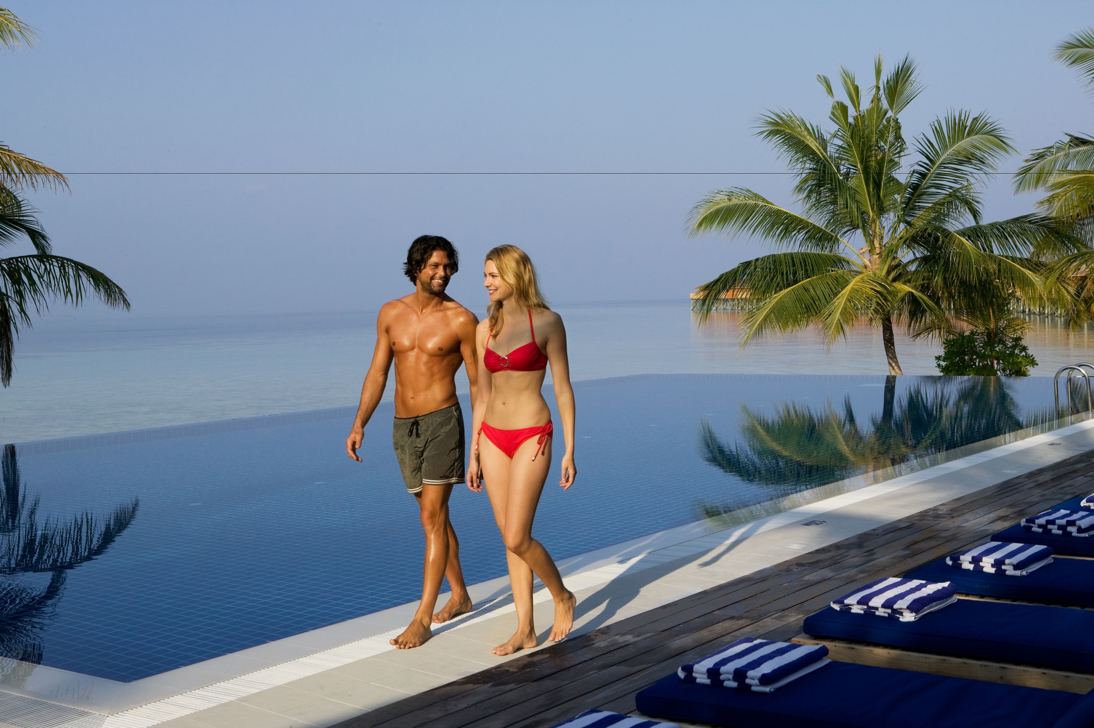 A couple walking on a poolside deck with a view of the ocean and palm trees.